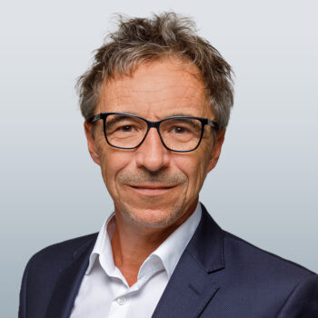 Ein Mann mittleren Alters mit kurzen, zerzausten Haaren und Brille, der einen marineblauen Blazer &uuml;ber einem wei&szlig;en Hemd tr&auml;gt und vor einem hellgrauen Hintergrund mit Farbverlauf leicht l&auml;chelt.
