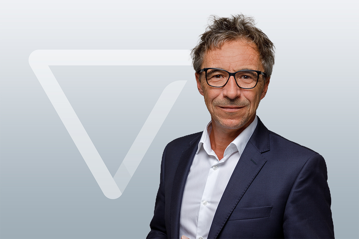 Ein Mann in einem marineblauen Anzug und mit Brille steht vor einem grauen Hintergrund mit einer weißen geometrischen Form und lächelt leicht in die Kamera.